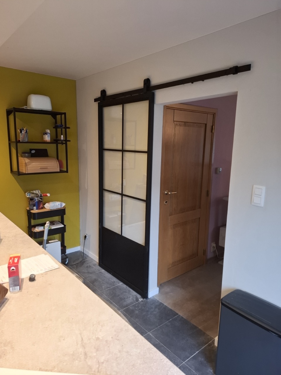 Zwarte stalen schuifdeur met glazen panelen naast een houten binnendeur in moderne woonkamer met gele muur en grijze tegelvloer